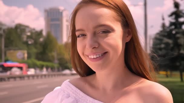 Porträtt av unga glada ingefära kvinna i vit skjorta ler mot kameran, urban street nära road i bakgrunden — Stockvideo