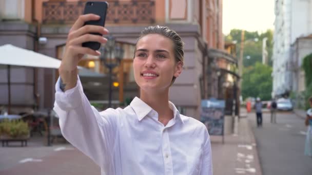 Современная женщина стоит на улице и делает селфи на смартфоне днем, улыбается, коммуникационная концепция, урбанистическая концепция — стоковое видео