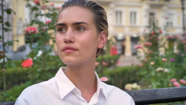 Jeune femme moderne est assis sur le banc et regarder à la caméra en journée, concept de détente, fond flou — Video