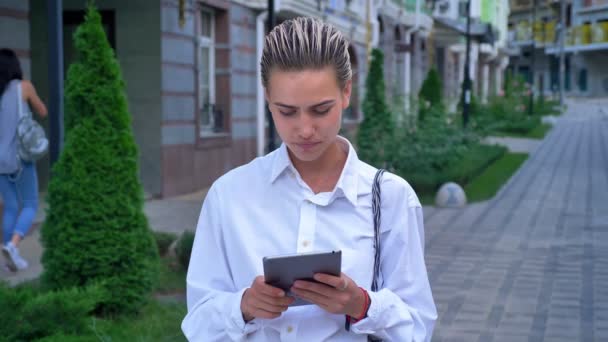 Junge moderne Frau steht auf der Straße und tippt auf Tablet, schaut vor der Kamera, Kommunikationskonzept, urbanes Konzept — Stockvideo