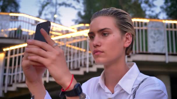 Jovem mulher moderna está de pé na ponte, tira foto do pôr do sol no smartphone, enviá-lo, conceito de comunicação, escada com luzes no fundo — Vídeo de Stock