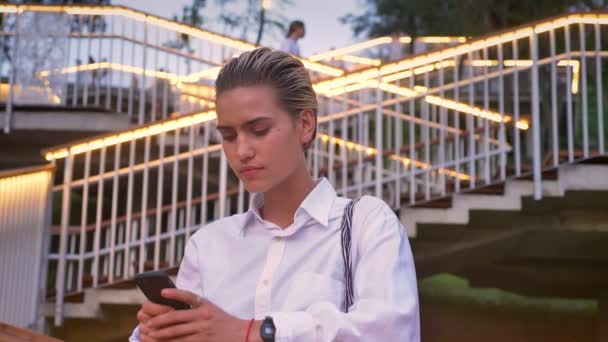 Bella donna moderna è in piedi sul ponte, guardando in smartphone, guardando la fotocamera, scala con luci sullo sfondo — Video Stock