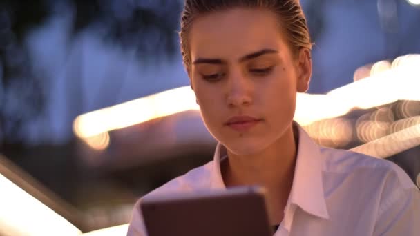 Mladá moderní žena sedí na žebříku, klepnutím na tabletu v večer, rozmazané světla na pozadí — Stock video