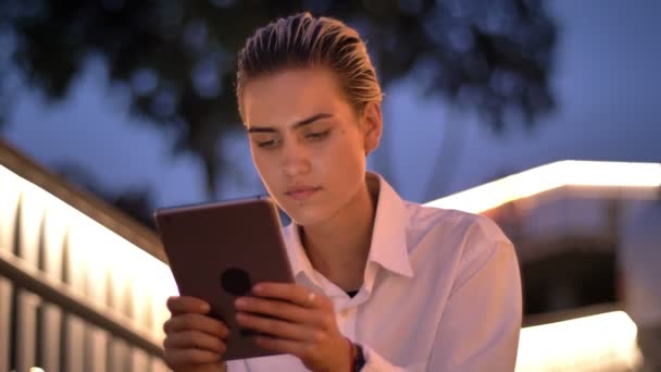 Giovane donna moderna è seduta sulla scala, toccando tablet in serata, sorridente, luci sfocate sullo sfondo — Video Stock