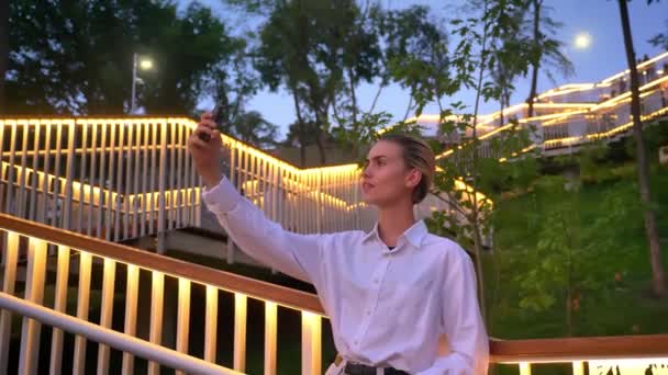 Сучасна жінка стоїть на драбині в парку, робить селфі на смартфоні ввечері, посміхається, концепція спілкування — стокове відео