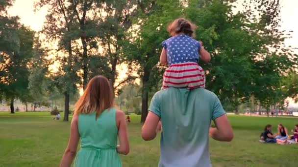 La silueta de la familia joven está caminando en el parque en verano, la chica se sienta sobre los hombros de los padres, puesta de sol — Vídeo de stock