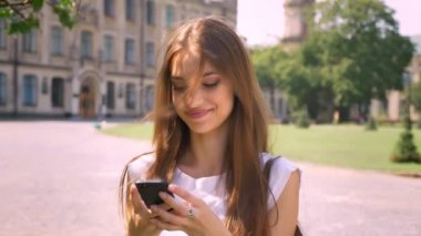 Seksi Bayan duruyor parkta gündüz de tipik haber smartphone, kameraya, izlerken gülümsüyor