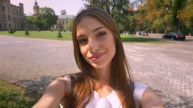 Güzel genç kadın duruyor parkta gündüz, kamera tutan, selfie, gülümseyen, iletişim kavramı yapar