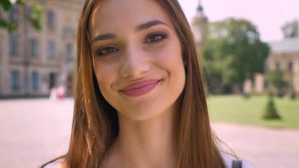 Nice young woman is standing in park in daytime, watching at camera, smiling, blurred background — Stock Video