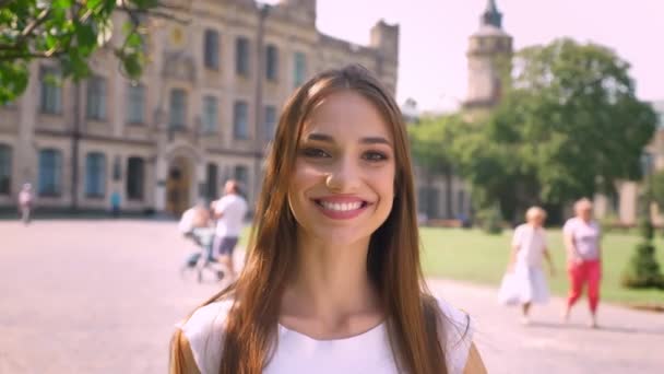 Nice young woman is standing in park in daytime, watching at camera, smiling, people walk on background — Stock Video