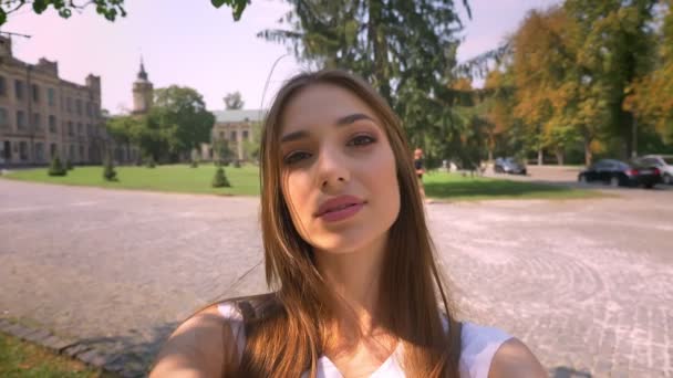 Jovem bonita está de pé no parque durante o dia, segura a câmera, faz selfie, conceito de comunicação — Vídeo de Stock