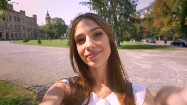 Belle jeune femme est debout dans le parc en journée, tient une caméra, fait du selfie, montre un signe de paix, concept de communication — Video
