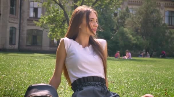 Seksowna Pani siedzi na trawie w parku w ciągu dnia, oglądając aparatu, uśmiechając się, zrelaksować się koncepcja — Wideo stockowe
