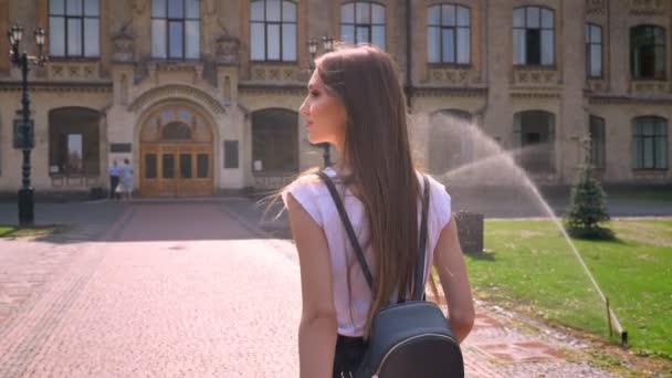 Nice young woman walks along in park in daytime in summer, turns head, building on background — Stock Video