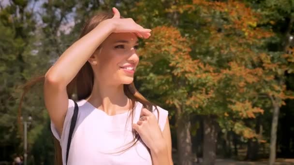 Mooie jonge vrouw squints, glimlachen, kruisen weg in park overdag in de zomer, kijken rond, ga naar Universiteit — Stockvideo