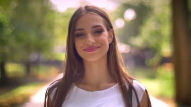 Güzel bir Bayan gündüz de izlerken kamera, gülümseme, bulanık backgrouns parkta duruyor