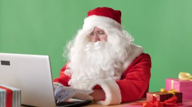 Ciddi Santa Claus oturma üstünde laptop çalışma, yazarak, seyir, kamera ve elini sallayarak, arka planda jest chromakey tebrik.
