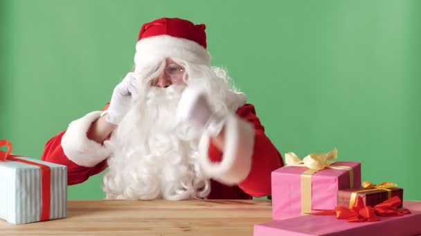 Onda Santa Claus talar på telefonen, slår näven på bord, sitter vid bord med gåvor, grön chromakey i bakgrunden. — Stockvideo