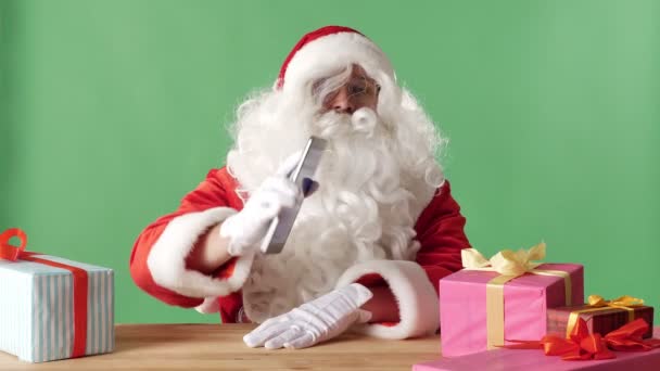 Smiling Santa Claus montrant tablette avec écran bleu à l'appareil photo, cadeaux sur la table, vert chromatique en arrière-plan . — Video