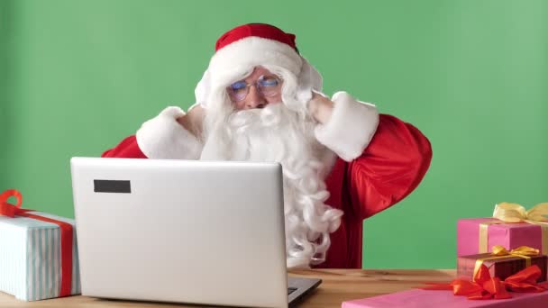 Santa Claus sonriente se sienta detrás de una computadora portátil escuchando música en los auriculares y bailando, cromakey verde en el fondo . — Vídeos de Stock