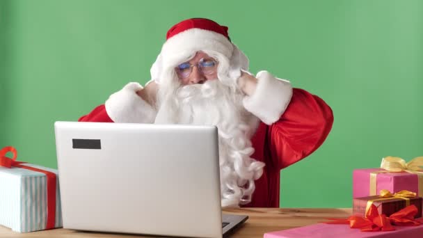 Babbo Natale sorridente siede dietro un computer portatile ascoltando musica su cuffie e ballando, cromakey verde sullo sfondo . — Video Stock