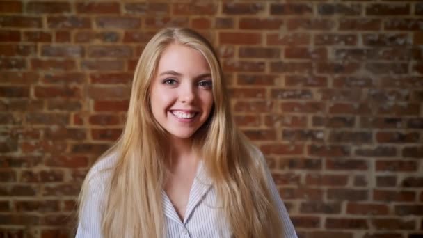 Joven chica rubia caucásica sonriendo y mirando a la cámara, retrato, pared de ladrillo en el fondo — Vídeos de Stock