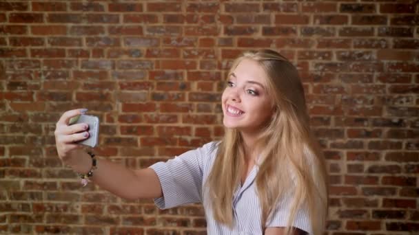 Νεαρή ξανθιά Καυκάσιος κάνοντας selfie, γέλιο, χαμόγελο, φωτογραφία, smartphone, πορτρέτο, τοίχο από τούβλα στο παρασκήνιο. — Αρχείο Βίντεο