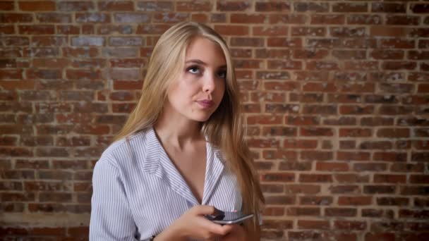Unga kaukasiska blondinen som använder en smartphone, maskinskrivning, svepa, porträtt, tegelvägg i bakgrunden — Stockvideo