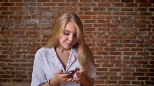 年轻的白种金发女郎使用智能手机显示智能手机屏幕, 蓝屏, 肖像, 砖墙背景 — 图库视频影像