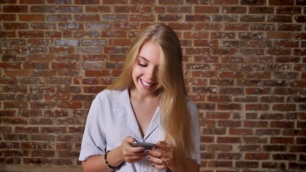 Joven rubia caucásica usando un teléfono inteligente que muestra una pantalla de teléfono inteligente, pantalla verde, retrato, pared de ladrillo en el fondo — Vídeos de Stock