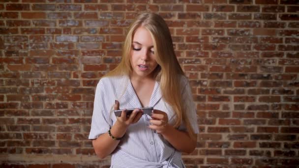 Jeune fille blonde caucasienne montrant écran de smartphone, écran vert, recommande l'application, comme signe, portrait, mur de briques en arrière-plan — Video