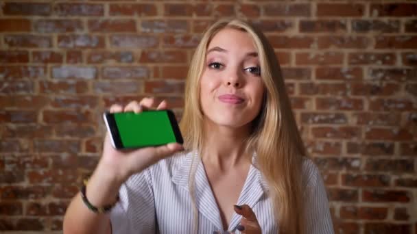 Giovane ragazza bionda caucasica che mostra lo schermo dello smartphone, schermo verde, raccomanda l'applicazione, come segno, ritratto, muro di mattoni in background — Video Stock