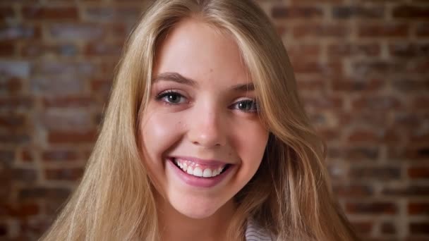 Young caucasian blonde girl smiling and looking at the camera, portrait, brick wall in the background — Stock Video