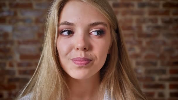 Jeune blonde caucasienne souriant, pensant, rêvant et regardant la caméra, portrait, mur de briques en arrière-plan — Video