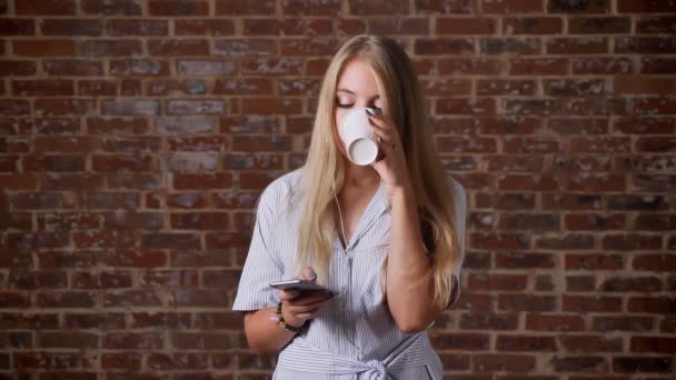 Joven rubia caucásica Escuchando música en un smartphone con auriculares, bailando, bebiendo café, retrato, pared de ladrillo en el fondo — Vídeos de Stock