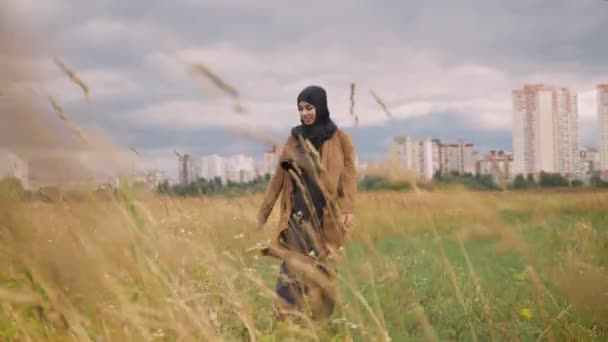 Jonge gelukkig Moslimvrouw in hijab tarweveld doorlopen en glimlachend, stad met wolkenkrabber in achtergrond — Stockvideo