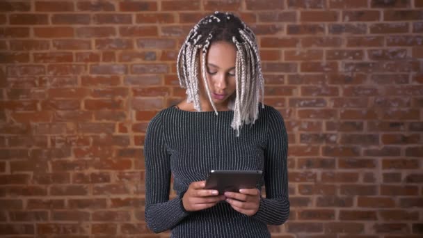 Bir tablet bilgisayar kullanarak, yazarak dreadlocks ile genç Afrikalı kız. Tuğla duvar içinde belgili tanımlık geçmiş. — Stok video