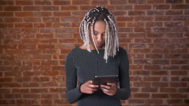 Chica africana joven con rastas usando una tableta, escribiendo. Muro de ladrillo en el fondo . — Vídeo de stock