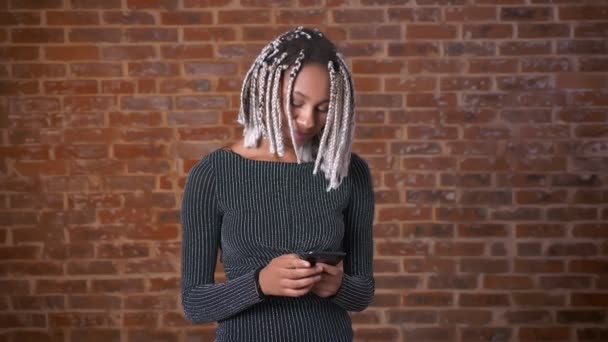Chica africana joven con rastas usando un teléfono inteligente, escribiendo. Muro de ladrillo en el fondo . — Vídeos de Stock