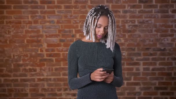 Jeune fille africaine avec dreadlocks à l'aide d'un smartphone regardant la caméra et souriant, Mur de brique en arrière-plan . — Video