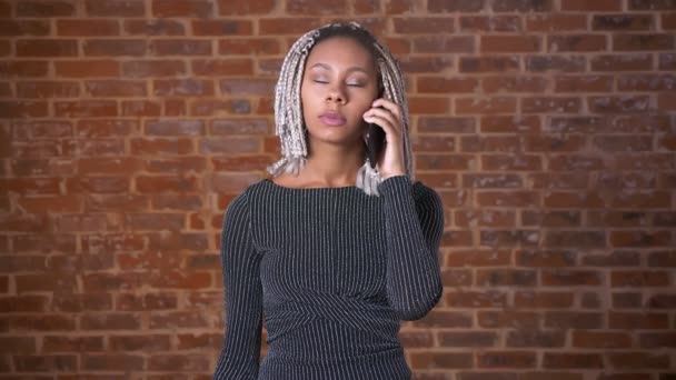 Afrikaanse meisje met dreadlocks praten aan de telefoon, bakstenen muur op de achtergrond. — Stockvideo