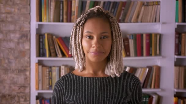 Jong Afrikaans meisje met dreadlocks glimlachen en kijken naar camera, boekenplanken op de achtergrond — Stockvideo