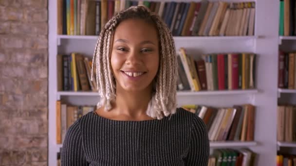 Jovem africana com dreadlocks sorrindo, rindo e olhando para a câmera, prateleiras do livro no fundo — Vídeo de Stock