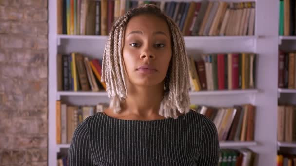 Joven chica seria africana con rastas chica mirando a la cámara, cerrando los ojos, estantes de libros en el fondo — Vídeos de Stock