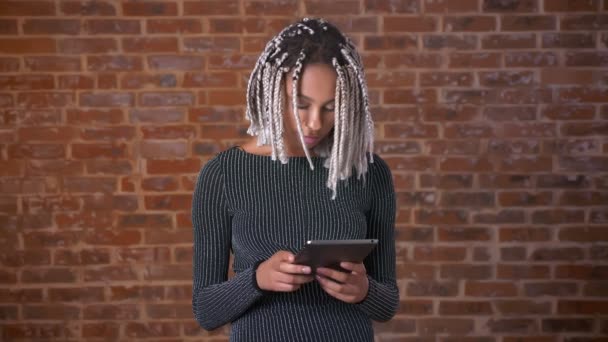 年轻的非洲女孩与辫子使用平板电脑看着相机和微笑, 墙上的背景. — 图库视频影像