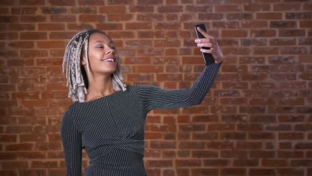 Selfie をやって、バック グラウンドでれんがの壁を笑顔のスマート フォンを使用してドレッドヘアを持つ若いアフリカの女の子. — ストック動画
