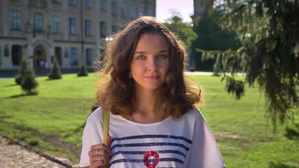 Portrét mladé usměvavé kavkazské bruneta v parku, univerzita v pozadí — Stock video