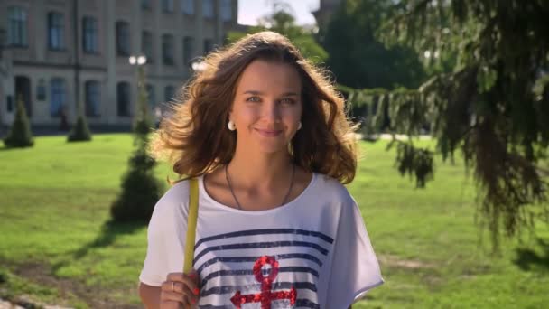 Retrato de una joven sonriente, riendo morena caucásica en el parque, universidad en el fondo — Vídeos de Stock