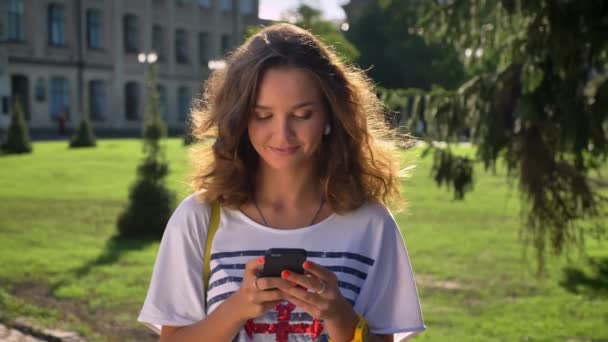 Joven chica caucásica está de pie en un parque y está utilizando un teléfono inteligente, mecanografía, universidad en el fondo — Vídeo de stock