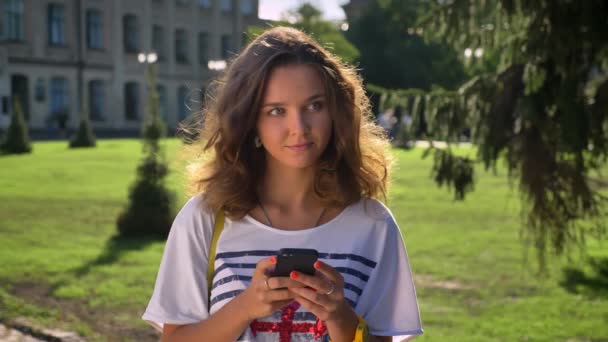 Молодая кавказская девушка стоит в парке и пользуется смартфоном, думая, университет на заднем плане — стоковое видео